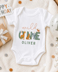A white onesie with the text "wild Oliver" printed on it, surrounded by various animal illustrations, is displayed on a bed with a "Happy Birthday" sign and confetti.