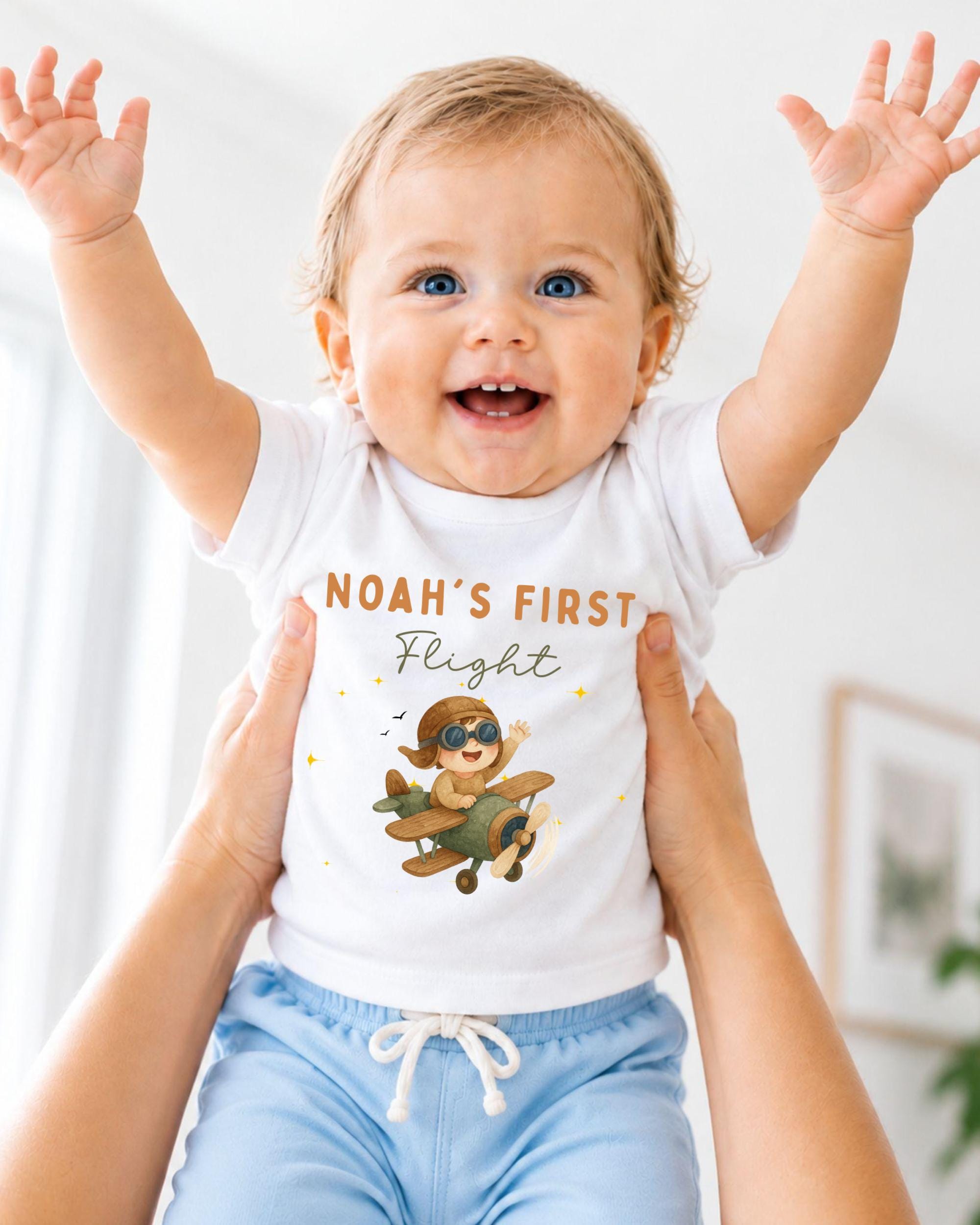 A smiling baby wearing a white t-shirt with a cartoon airplane and the text "Noah's first flight" is being held up by two people.