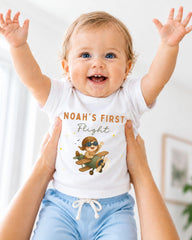 A smiling baby wearing a white t-shirt with a cartoon airplane and the text "Noah's first flight" is being held up by two people.