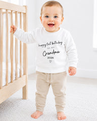 A smiling baby boy wearing a white sweatshirt with text that says "happy first birthday as my Grandma" and "2026" printed on it, standing next to a wooden crib.