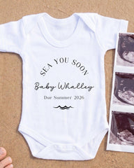 A white onesie with text and graphics printed on it, placed on a sandy surface next to a photograph of an ultrasound image.