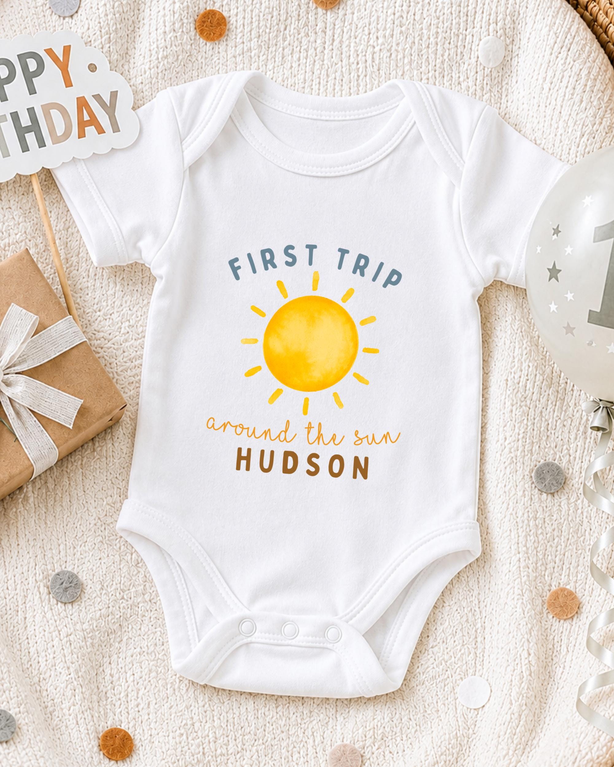 A white onesie with a sun graphic and the text "First Trip around the sun Hudson" printed on it, along with a "Happy Birthday" sign and a balloon with the number "1" on it, all placed on a white blanket with scattered confetti.