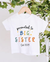 A white t-shirt with text that says "promoted to BIG SISTER Est 2029" printed on it.