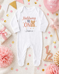 A white onesie with the text "Halfway to one" and a teddy bear graphic, surrounded by confetti, a pink balloon, and other baby-related items.