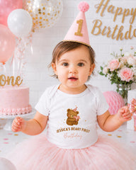 A young girl wearing a birthday hat and a white t-shirt with a teddy bear design is posing in front of a birthday cake and balloons.