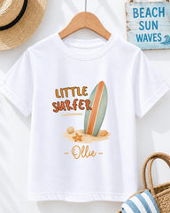 A white t-shirt with a graphic design of a surfboard and beach elements, along with the text "LITTLE SURFER - OLLIE" printed on it. The t-shirt is hanging on a wooden hanger, and there is a straw hat and a beach sign visible in the background.