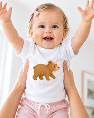 A smiling baby girl wearing a white t-shirt with a bear graphic and the name "Emma" printed on it, with her arms raised in a joyful gesture.