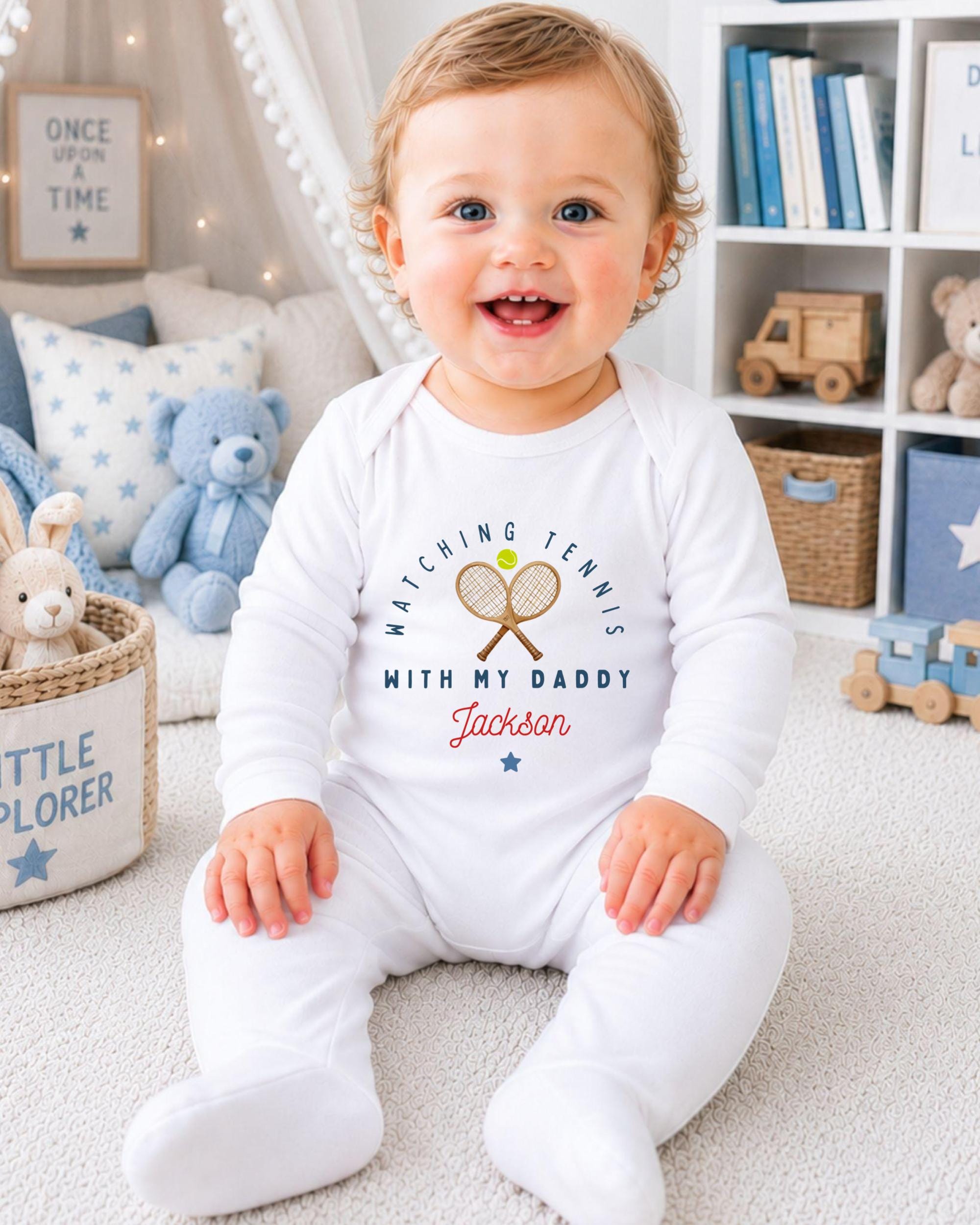A smiling baby boy wearing a white onesie with a tennis racket and the text "Matching Tennis with my Daddy Jackson" is sitting on the floor in a room with a bookshelf and stuffed animals in the background.