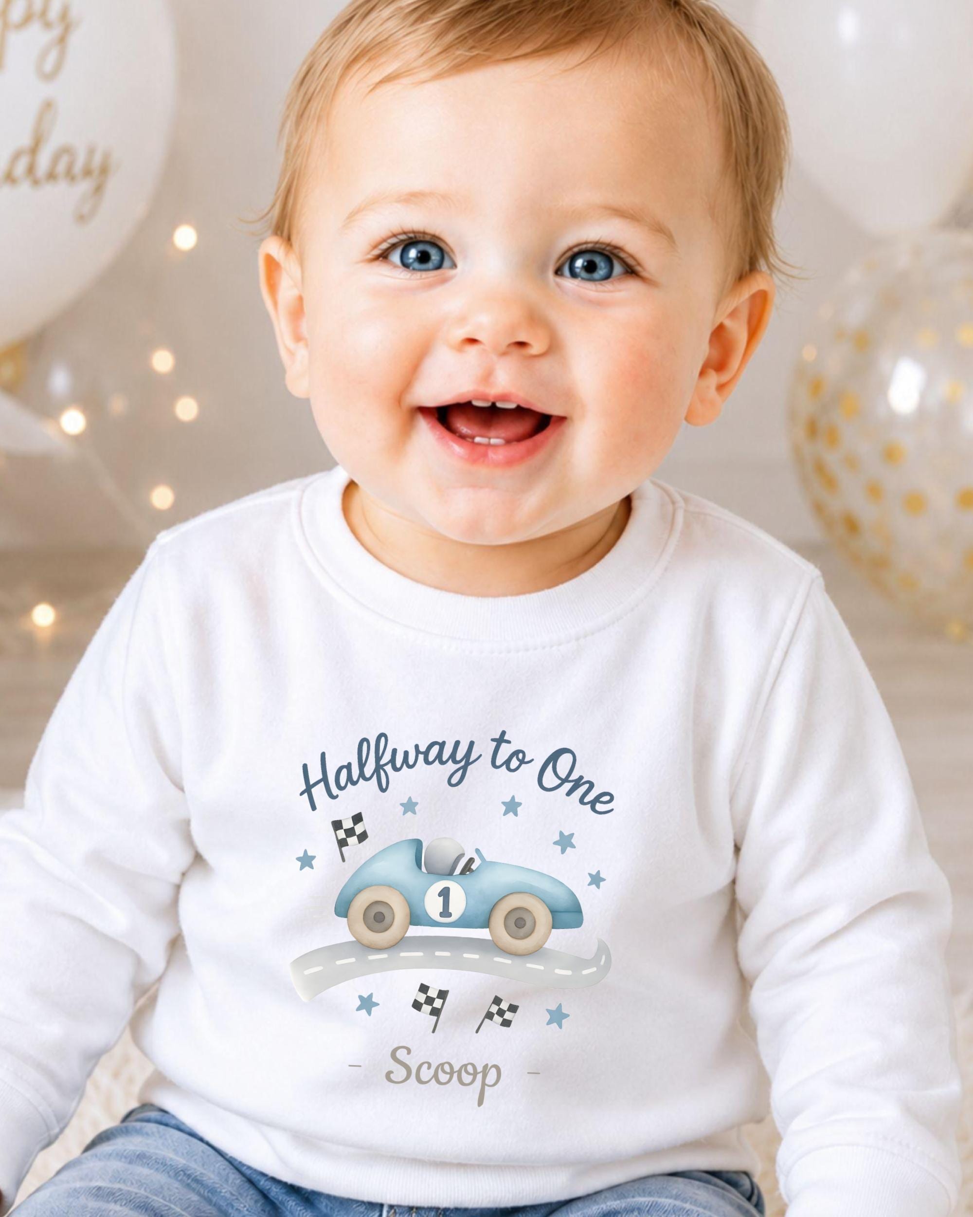 A smiling baby boy wearing a white shirt with a graphic of a race car and the text "Halfway to One Scoop" printed on it.