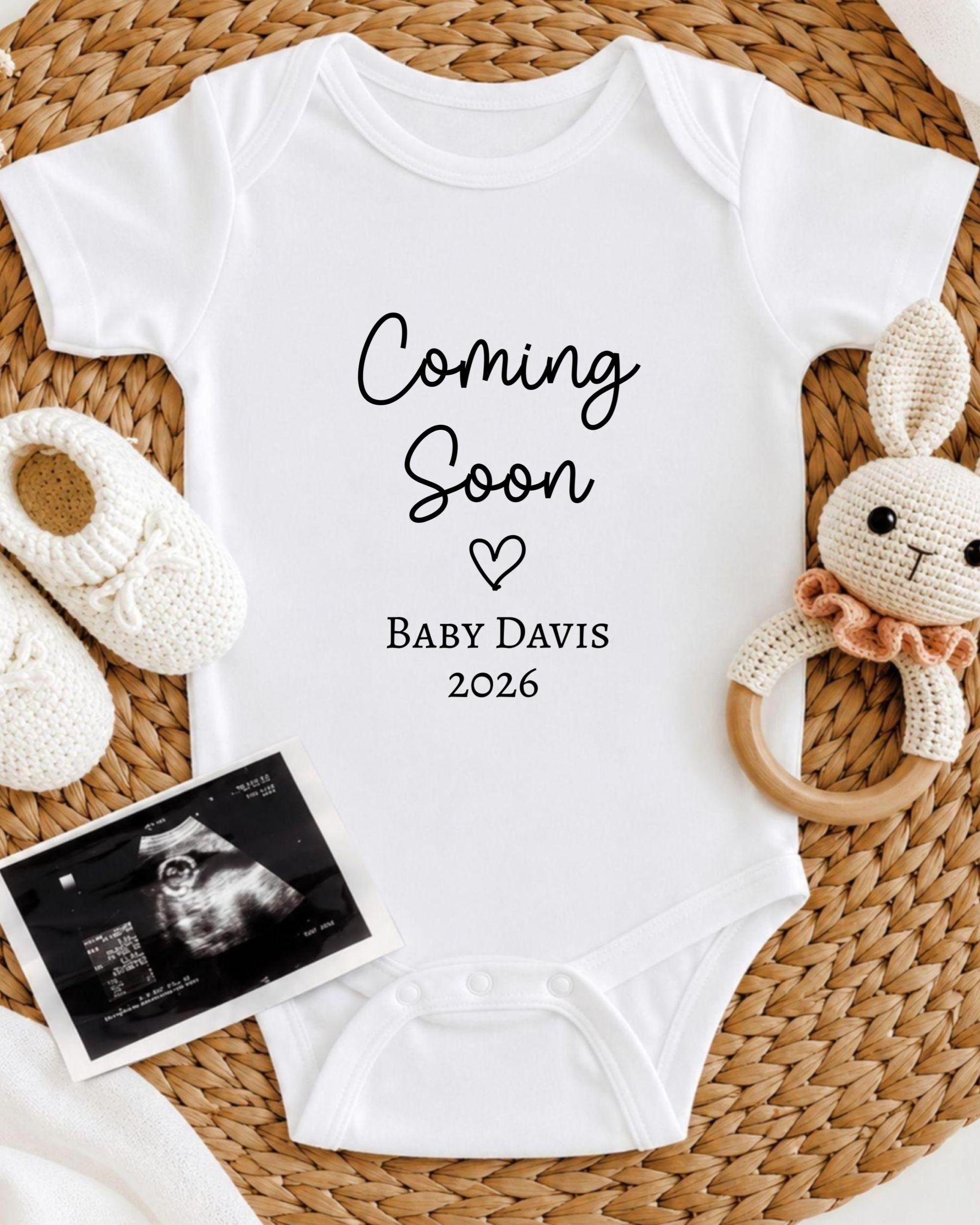 A white baby onesie with the text "Coming Soon Baby Davis 2026" printed on it, along with a crocheted bunny toy and a black and white photograph of an infant's head.