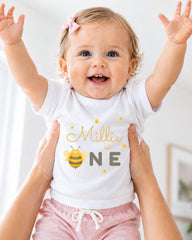 A smiling baby girl wearing a white t-shirt with a bee graphic and the text "Milly is one" is being held up by someone's arms.