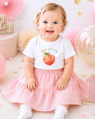 A smiling baby girl wearing a white t-shirt with an apple graphic and a pink skirt, sitting on a pink and white background with balloons and confetti.