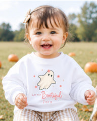A smiling baby girl wearing a white sweater with a ghost and flower design, sitting in a field with pumpkins in the background.
