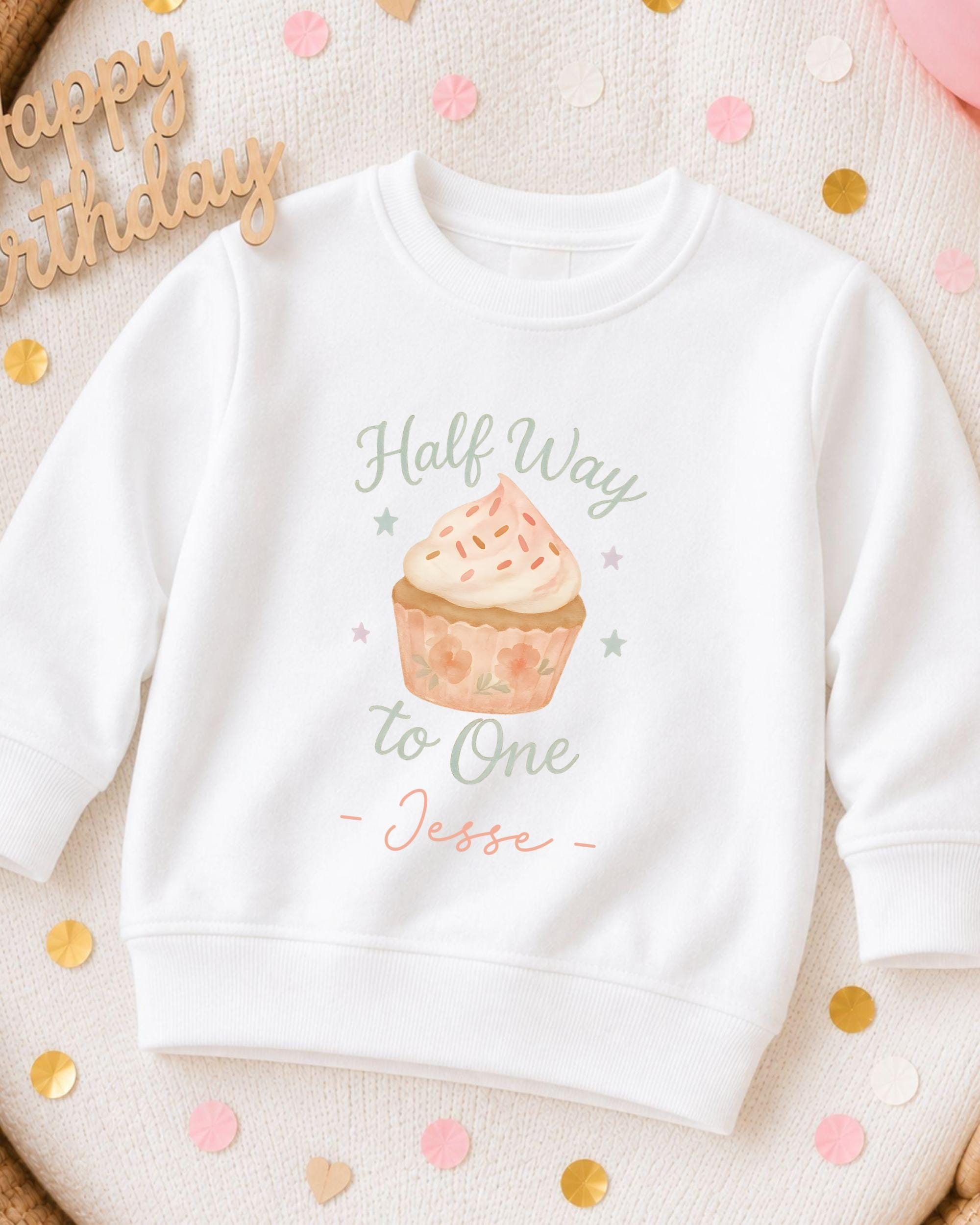 A white sweatshirt with a graphic of a cupcake and the text "Half Way to One Jesse" printed on it, surrounded by confetti.