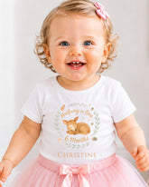 A smiling baby girl wearing a white t-shirt with a deer graphic and the text "Halfway to One 6 Months Christine" printed on it.
