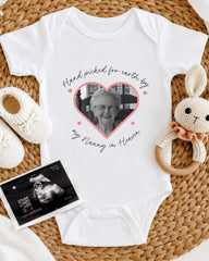 A white baby onesie with a heart-shaped photo of an elderly woman, surrounded by baby booties and a crocheted rabbit toy.