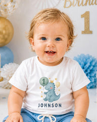 A smiling baby boy wearing a white t-shirt with a dinosaur graphic and the name "Johnson" printed on it, sitting in front of a birthday party decoration.