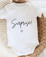A white baby onesie with the word "Surprise!" printed on it, placed on a woven basket.