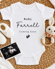 A white baby onesie with the text "Baby Farrell Coming Soon" printed on it, along with a pair of white crocheted shoes and a baby rattle. There is also an image of an ultrasound scan of a baby's belly.