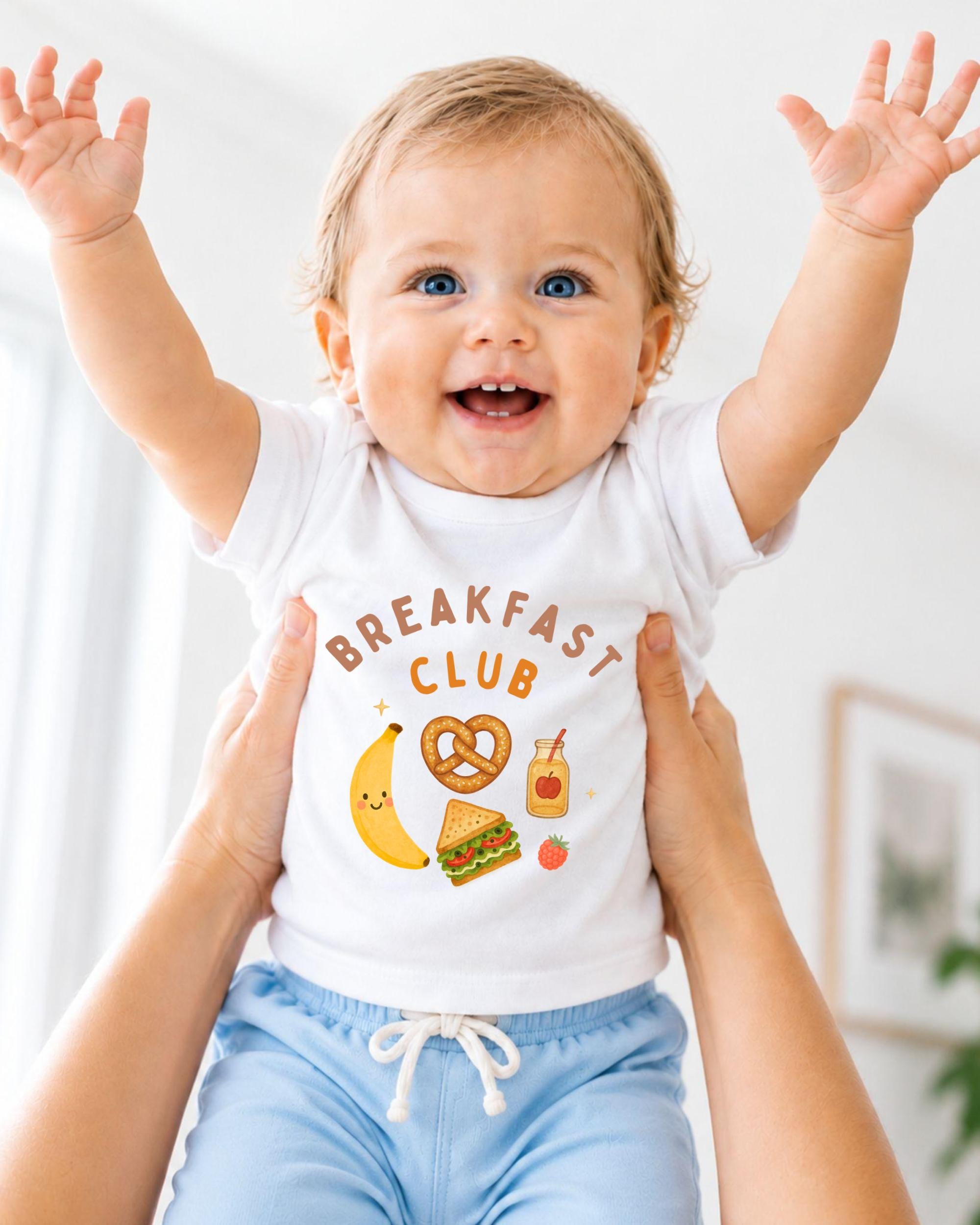 A smiling baby wearing a white shirt with the text "BREAKFAST CLUB" and various cartoon food items, such as a banana, pretzel, and sandwich, is being held up by two people.