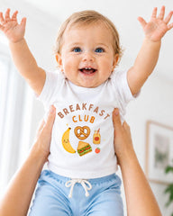 A smiling baby wearing a white shirt with the text "BREAKFAST CLUB" and various cartoon food items, such as a banana, pretzel, and sandwich, is being held up by two people.