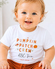A smiling baby wearing a white t-shirt with text and illustrations related to a pumpkin patch crew.