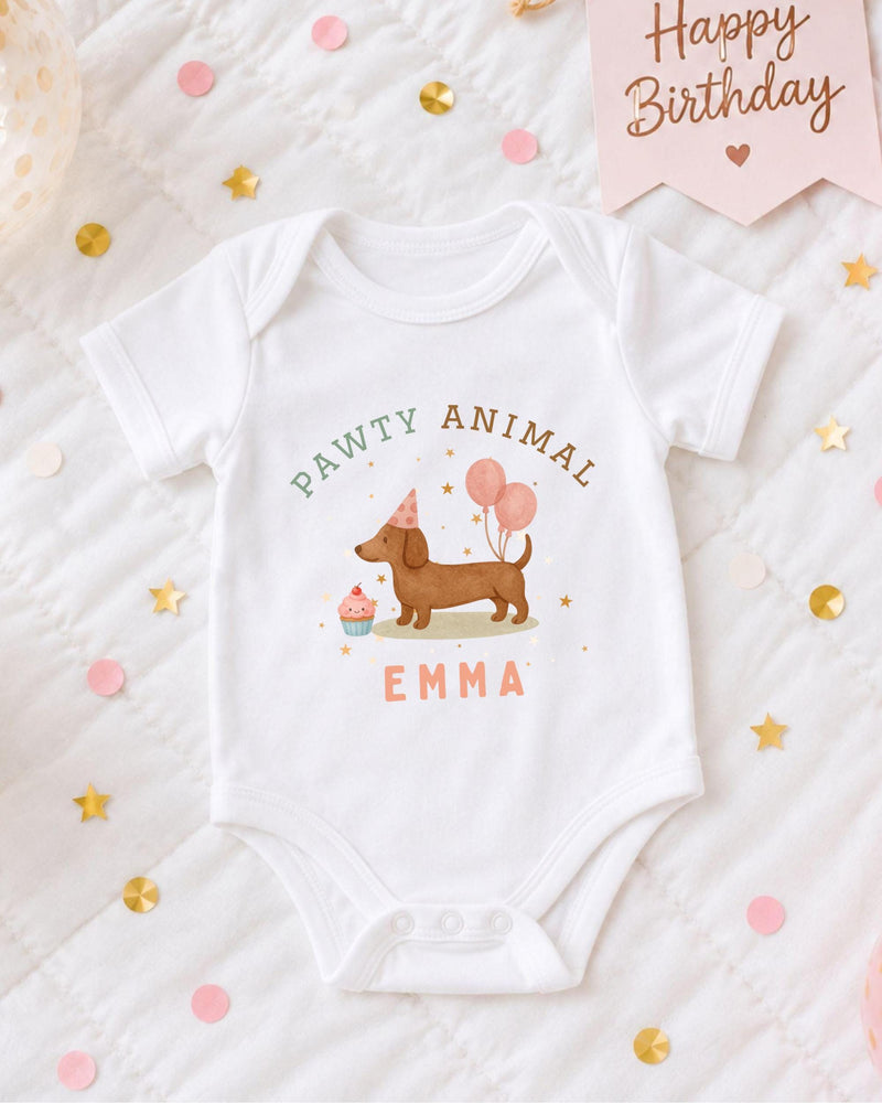 A white baby bodysuit with a dog and birthday party decorations, including a pink banner that says "Happy Birthday" and confetti.