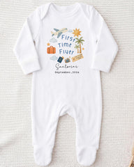 A white onesie with a graphic design on it, featuring a plane, a sun, a palm tree, and various travel-related icons.