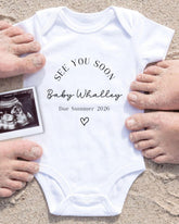a white baby onesie with the text "See you soon Baby Whaley Due Summer 2026" printed on it, surrounded by sand and a small black and white photograph of a baby's head.