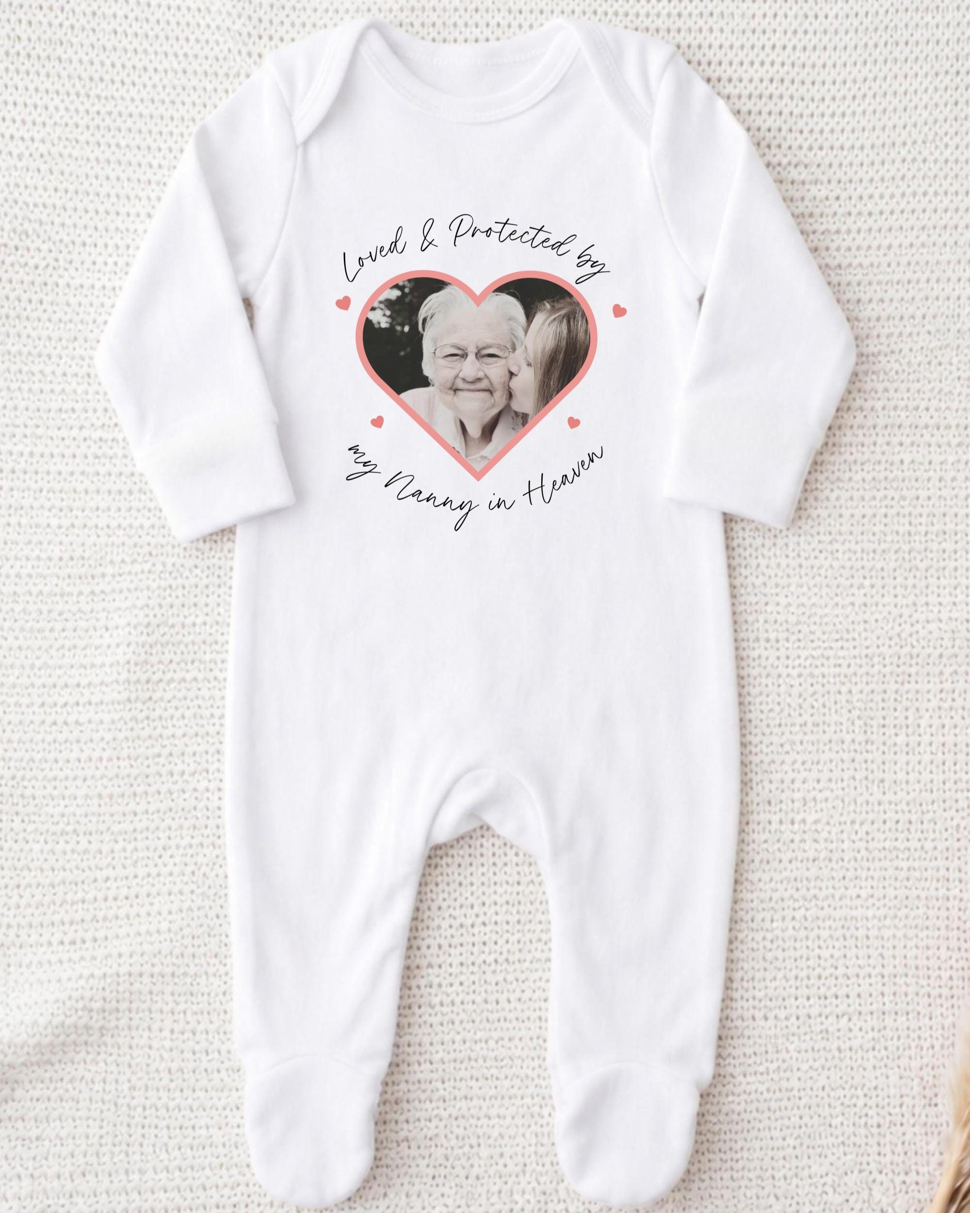 A white onesie with a heart-shaped photo of an elderly woman and the text "Loved & Protected by my Nanny in Heaven" printed on it.