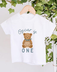 A white t-shirt with a graphic of a teddy bear and the text "Oscar is ONE" printed on it, hanging on a wooden hanger.