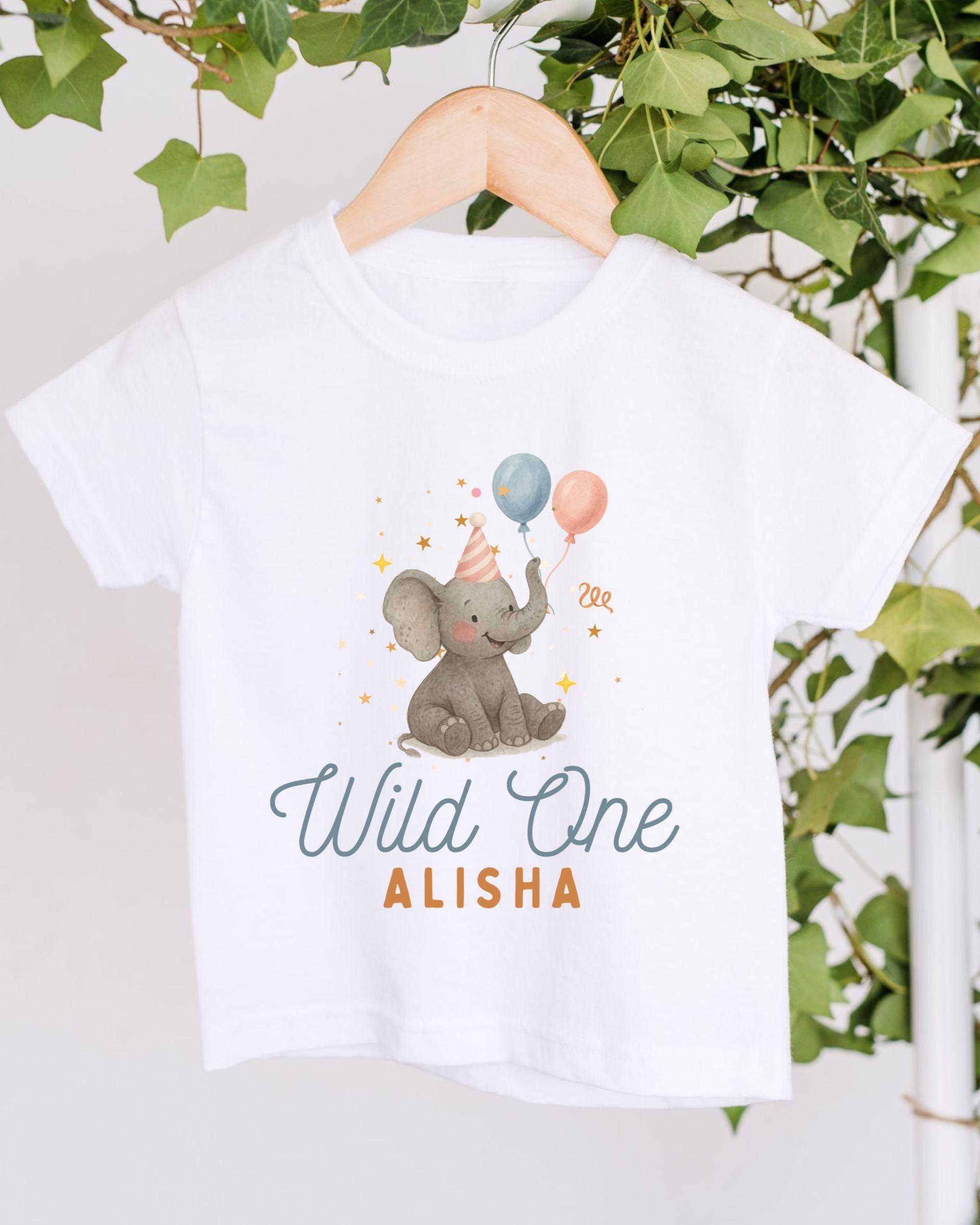 A white t-shirt with a graphic design of an elephant holding balloons and wearing a party hat, along with the text "Wild One Alisha" printed on it.