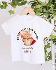 A white t-shirt with a graphic design featuring a bear wearing a red cape and a birthday hat, along with the text "Happy Birthday Daddy" and "love you lots Alfie".