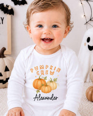 A smiling baby boy wearing a white onesie with a pumpkin and leaf design, sitting in front of Halloween decorations.