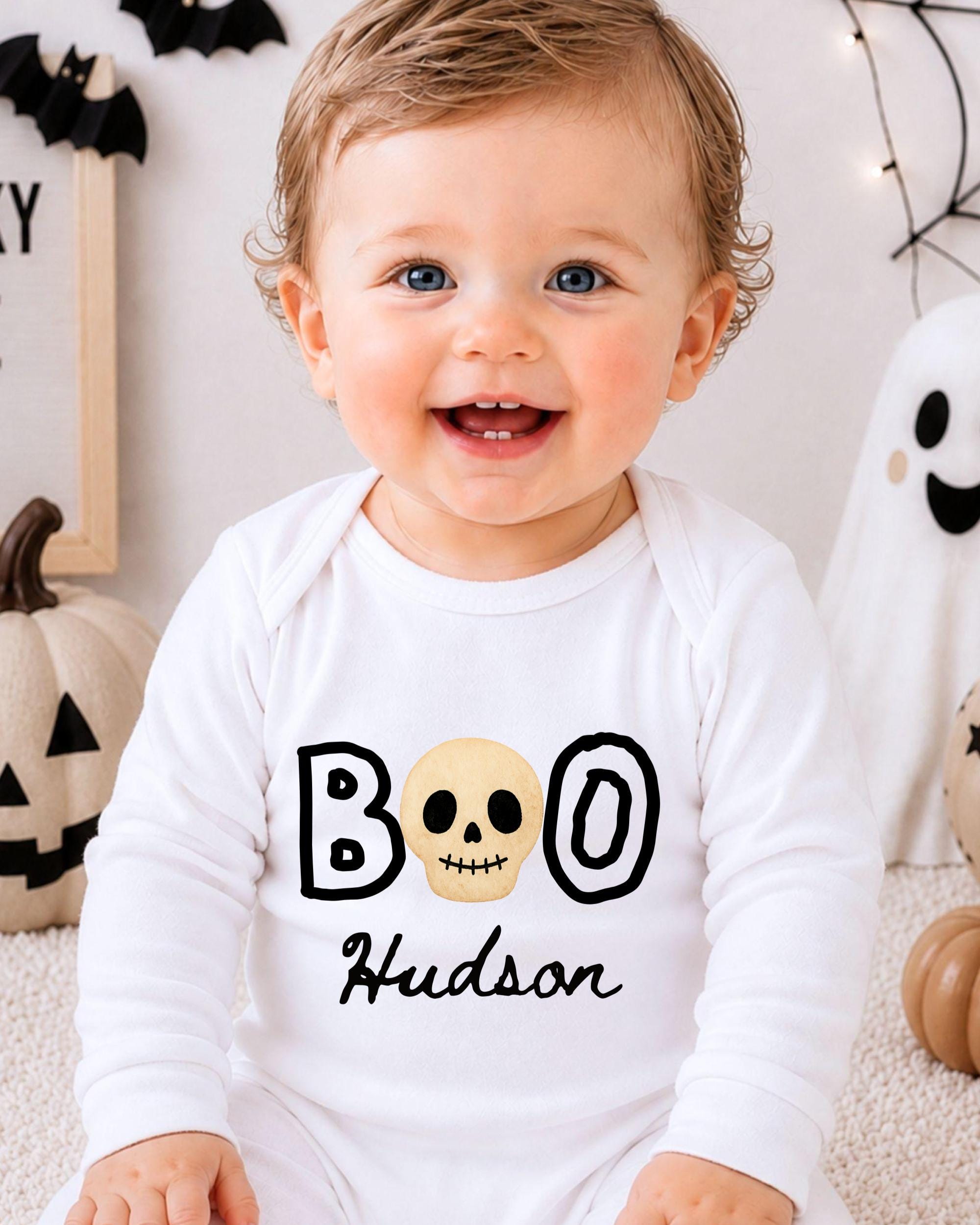 A smiling baby boy wearing a white onesie with the text "BOO Hudson" printed on it, sitting in front of Halloween decorations.