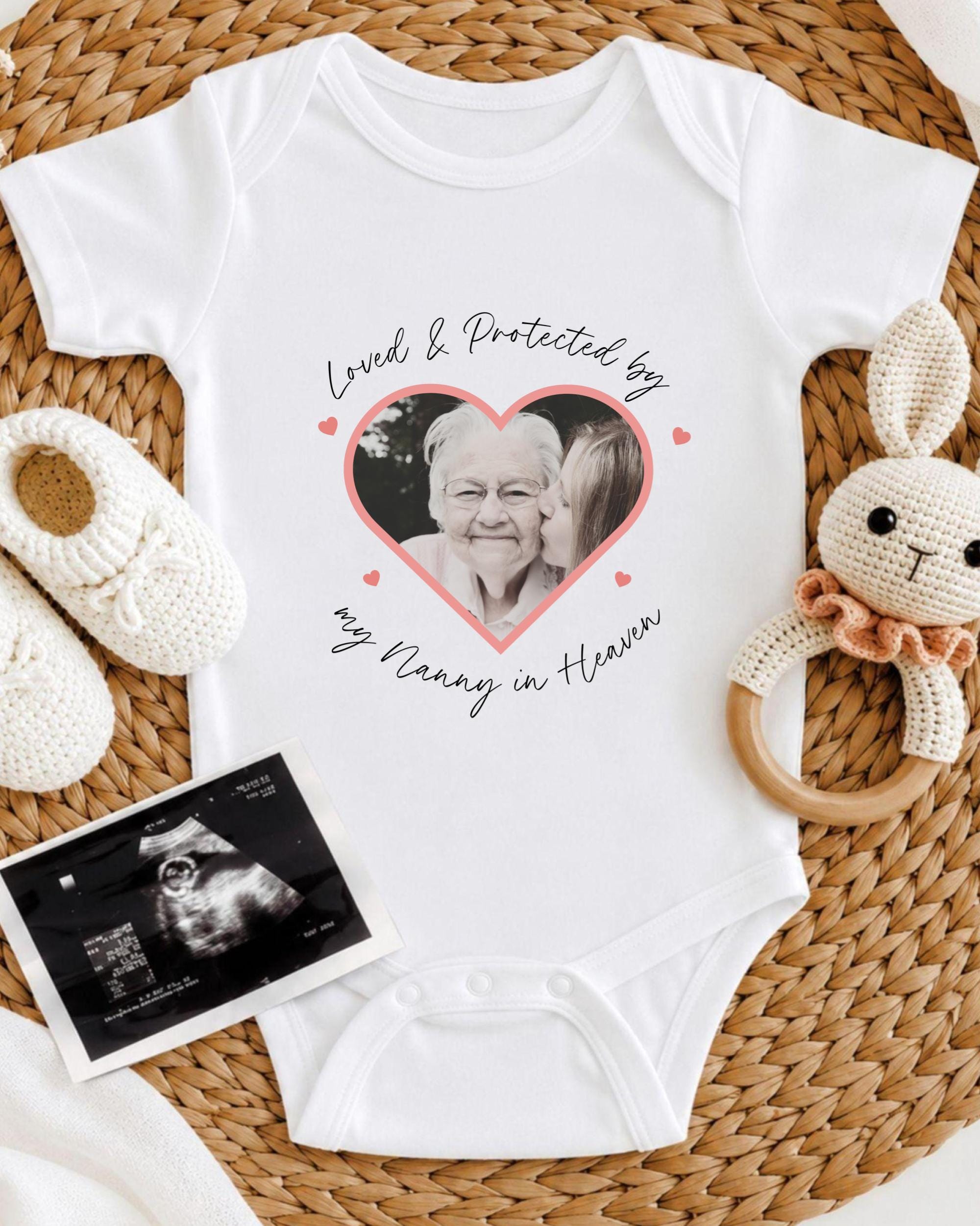 A white baby onesie with a heart-shaped photo of an elderly woman and the text "Loved & Protected by my Nanny in Heaven" printed on it, along with a pair of white crocheted baby booties and a black and white photograph of an infant's head.