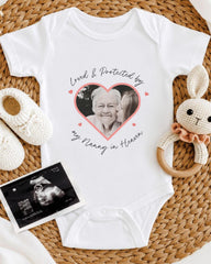 A white baby onesie with a heart-shaped photo of an elderly woman and the text "Loved & Protected by my Nanny in Heaven" printed on it, along with a pair of white crocheted baby booties and a black and white photograph of an infant's head.