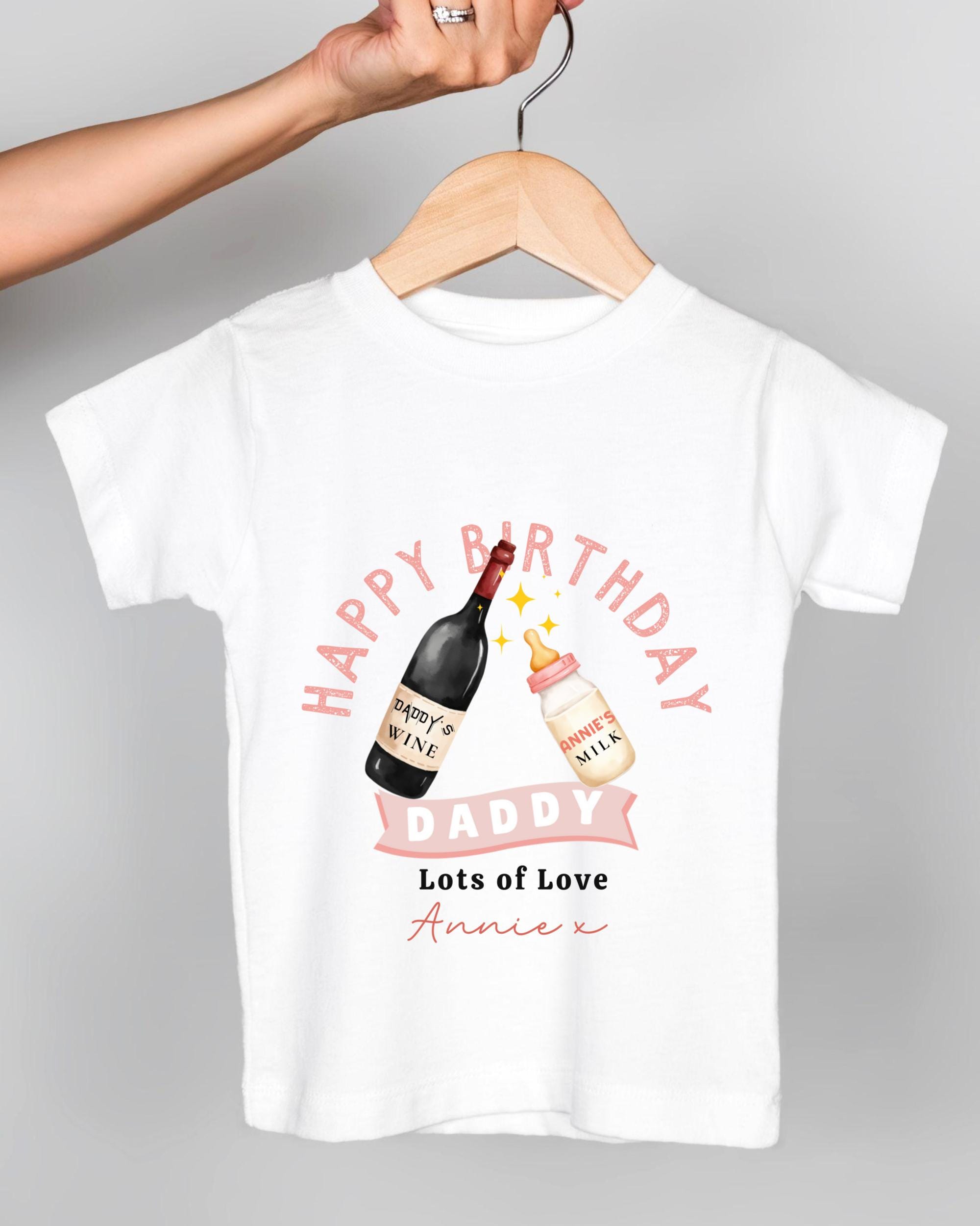 A white t-shirt with a graphic design featuring a bottle of wine and a baby bottle, along with the text "Happy Birthday Daddy Lots of Love Annie" printed on it.
