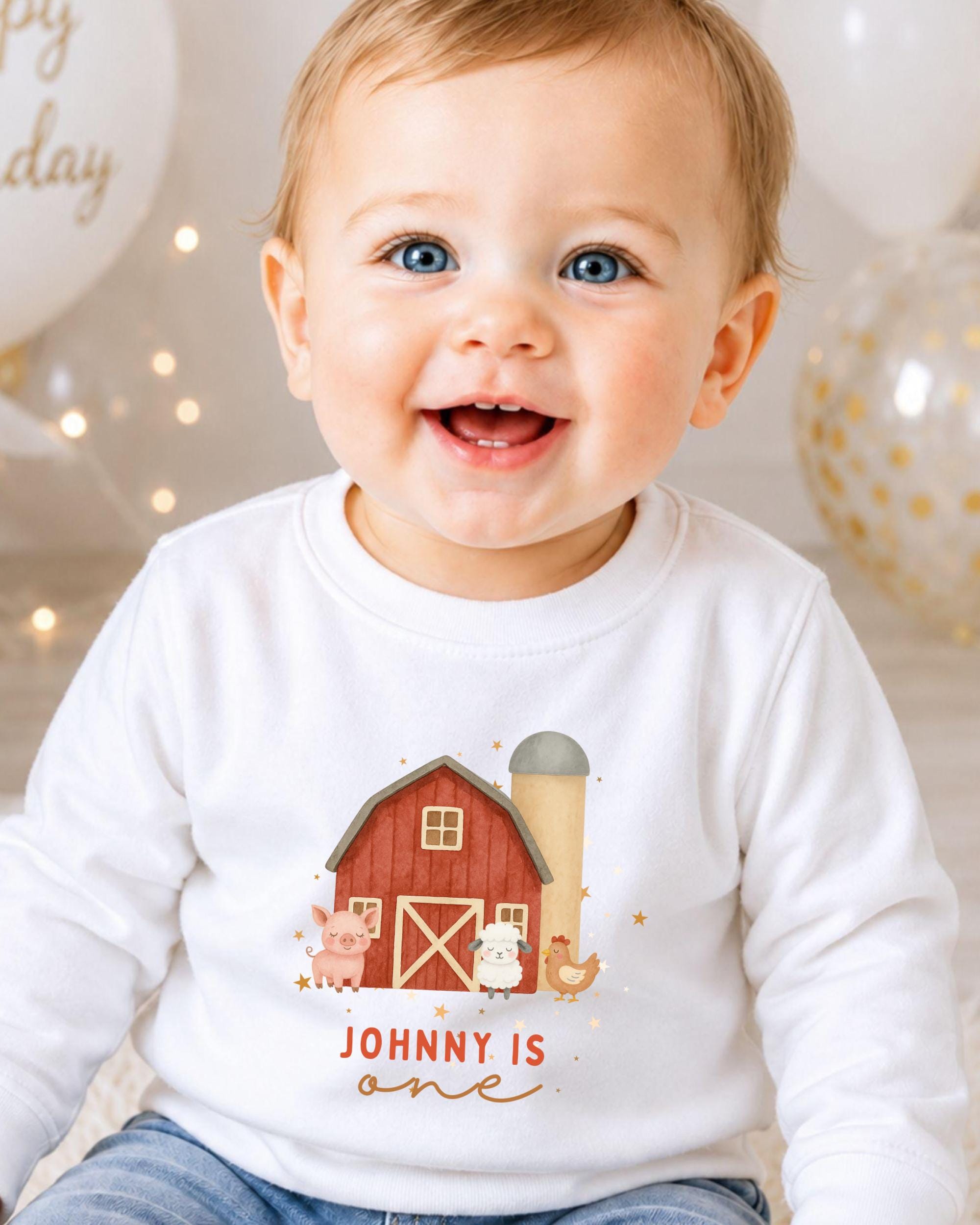 A smiling baby boy wearing a white shirt with a farmhouse graphic and the text "Johny is one" is sitting in front of birthday balloons.