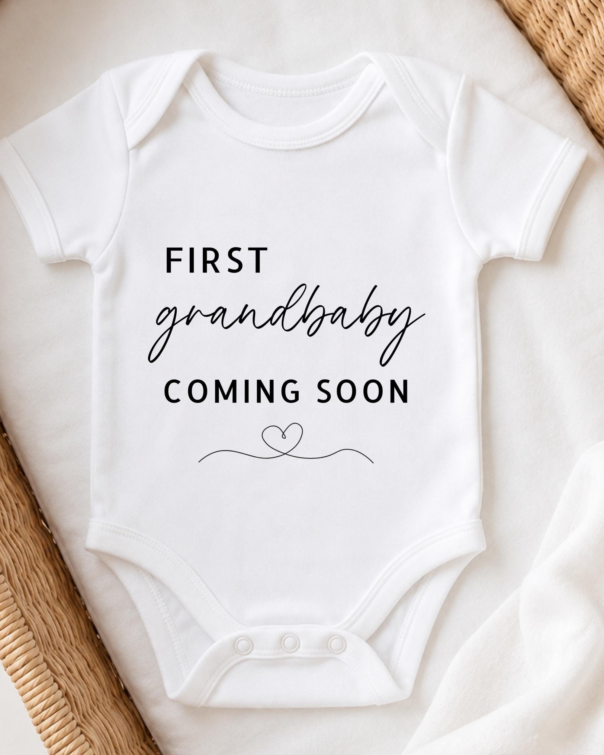 A white onesie with the text "FIRST GRANDBABY COMING SOON" printed on it, placed on a white surface.