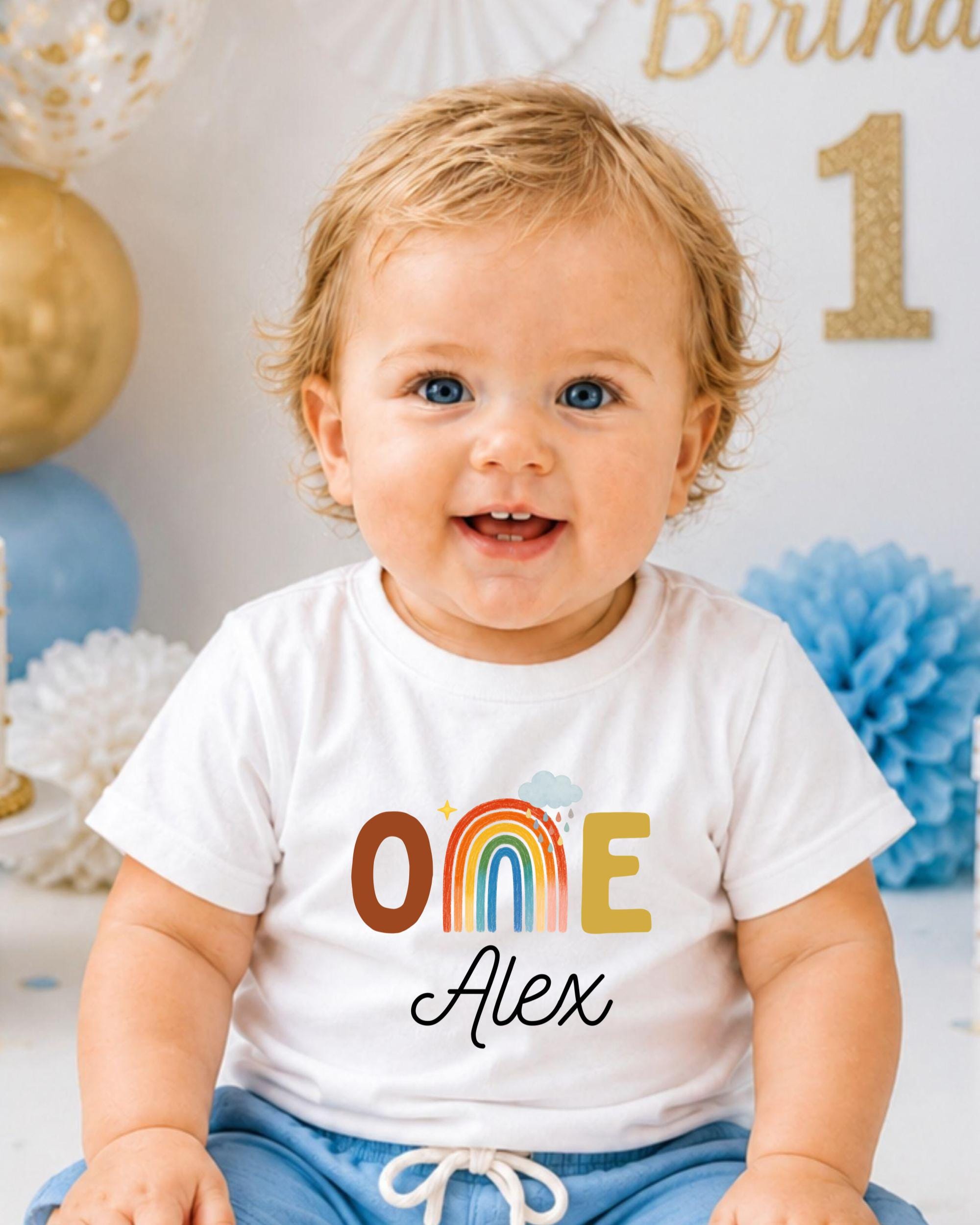 A smiling baby boy wearing a white t-shirt with a rainbow graphic and the name "Alex" printed on it, sitting in front of a birthday party backdrop.
