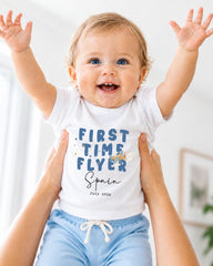 A smiling baby wearing a white t-shirt with the text "FIRST TIME FLYER Spain" and blue shorts, being held up by someone's arms.