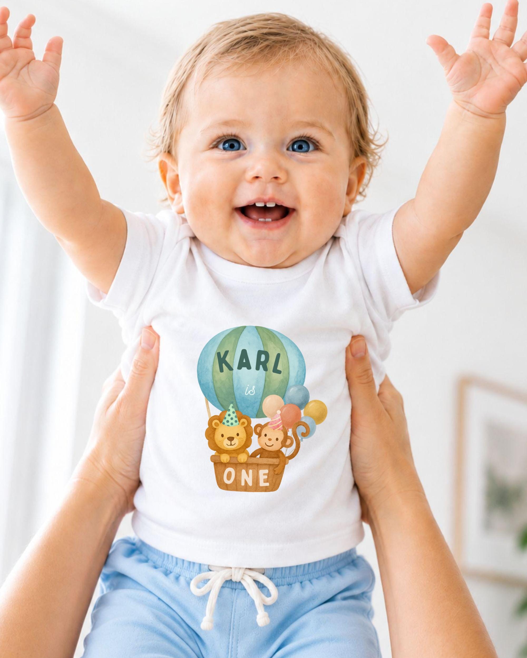 A smiling baby wearing a white shirt with a cartoon image of a hot air balloon and the name "Karl" printed on it, being held up by someone's arms.