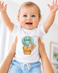 A smiling baby wearing a white shirt with a cartoon image of a hot air balloon and the name "Karl" printed on it, being held up by someone's arms.