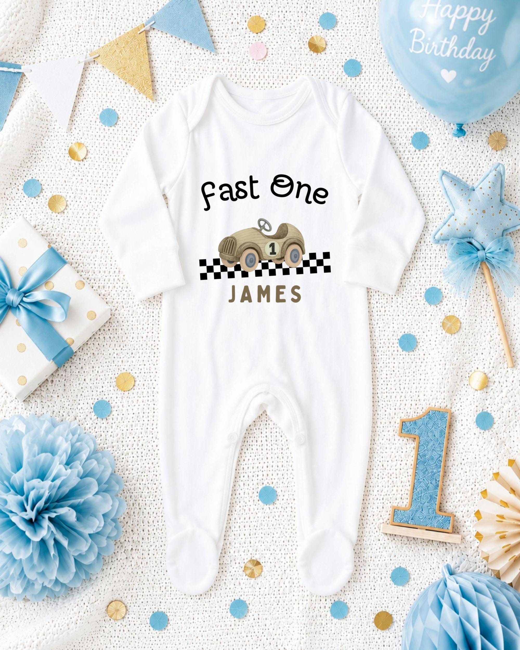 A white onesie with a race car design and the text "Fast One James" printed on it, surrounded by birthday decorations and confetti.