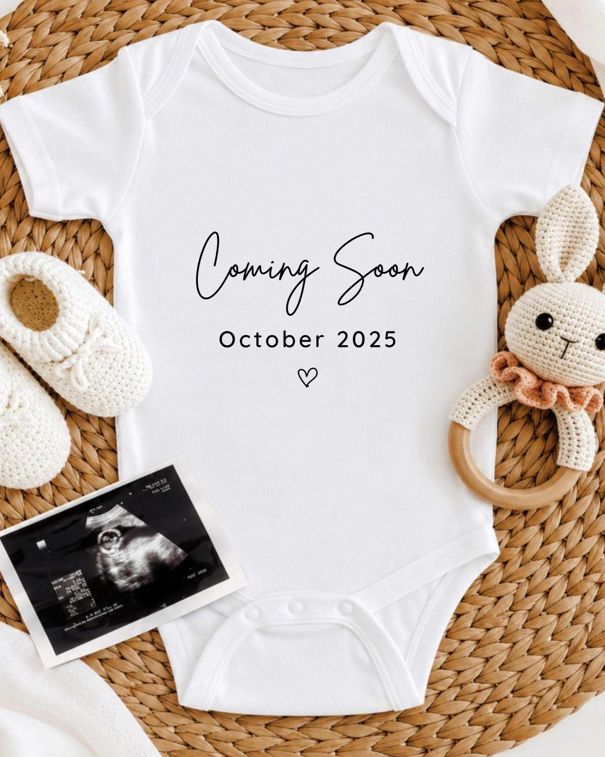 A white baby onesie with the text "Coming Soon October 2025" printed on it, along with a crocheted bunny toy and a black and white photograph of an infant's head.