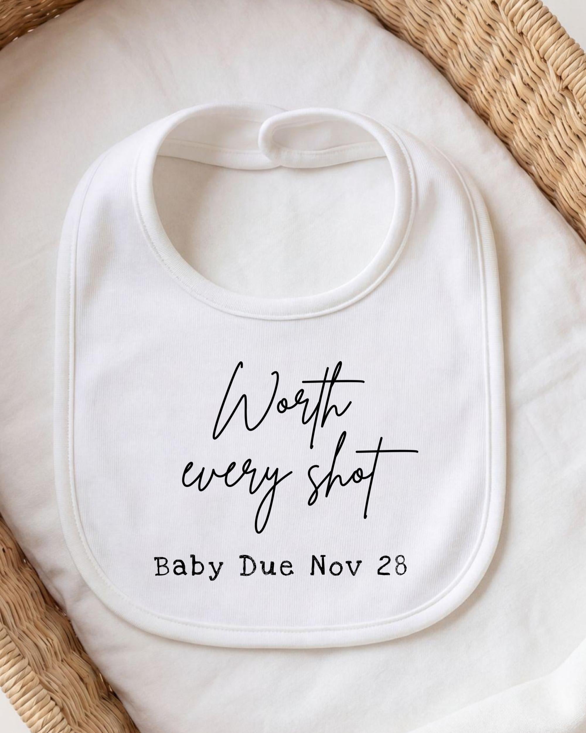A white bib with the text " Worth every shot Baby Due Nov 28" printed on it.