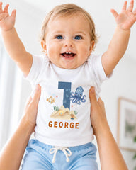 A smiling baby wearing a white shirt with a blue number 1 and an underwater-themed design, being held up by someone's arms.