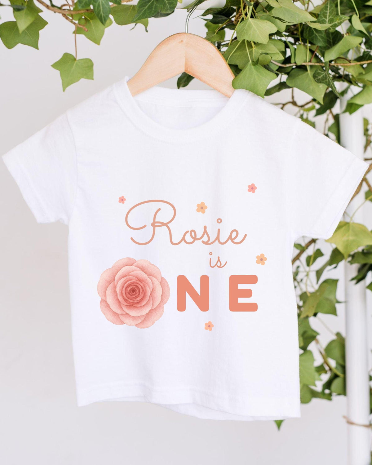 A white t-shirt with a rose design and the text "Rosa is ONE" printed on it, hanging on a wooden hanger.