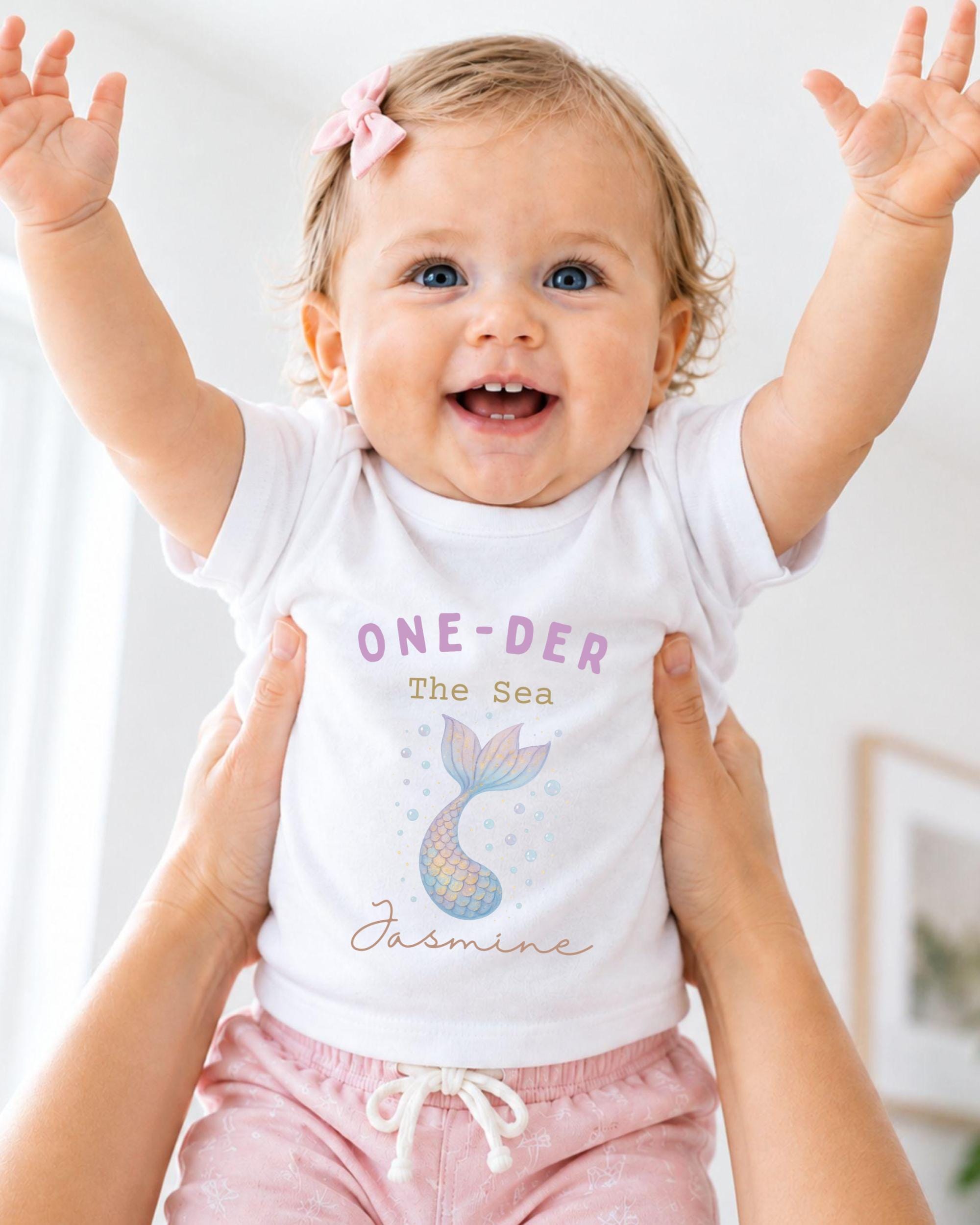 A smiling baby girl wearing a white t-shirt with a mermaid design and the text "One-DER The Sea Jasmine" is being held up by someone's arms.